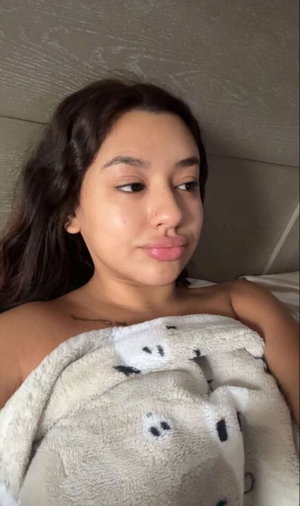 Woman with dark hair lying in bed, covered by a patterned blanket, showing shoulders and neck.