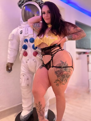 Curvy tattooed woman in gold top and black lingerie indoors.