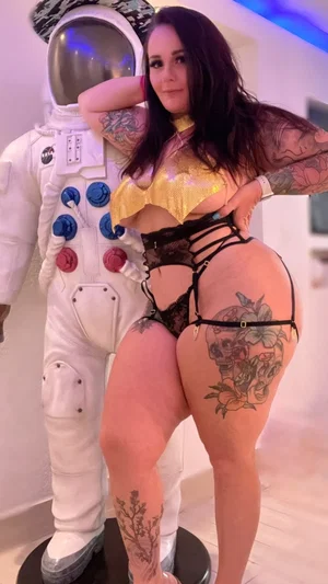 Woman with tattoos in gold top and black lace lingerie posing beside astronaut mannequin indoors.