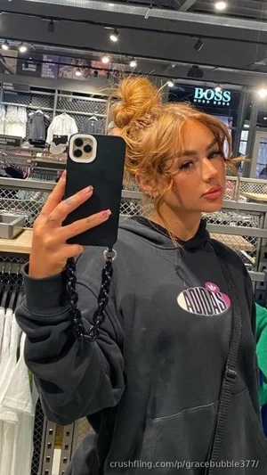 Woman with blonde hair in bun, wearing black hoodie, taking mirror selfie in clothing store.