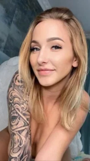Blonde woman with arm tattoos smiles in bathroom selfie.