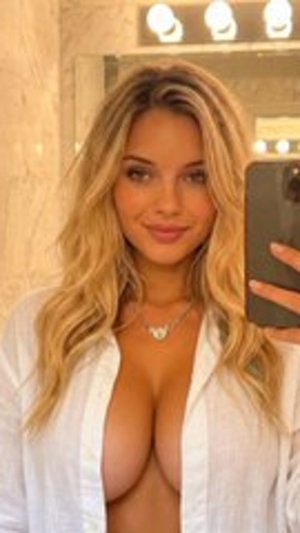 Woman with blonde hair in white shirt, bare chest, taking mirror selfie in bathroom.