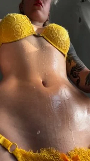Woman in yellow lace lingerie with wet skin and arm tattoo, close-up torso shot.