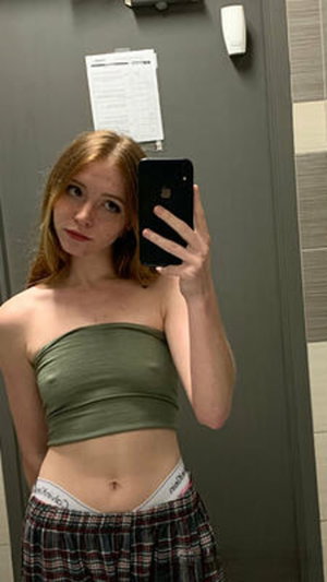 Red-haired woman taking mirror selfie in bathroom wearing green top.