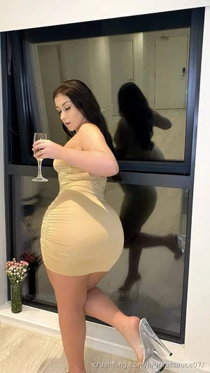 Woman in beige bodycon dress holding wine glass, standing by window with reflection.