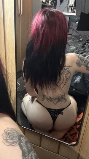 Woman with red-tipped hair and tattoos in black thong, kneeling in mirror selfie in bedroom.