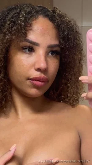 Woman with curly hair, bare chest, holding pink phone in bathroom mirror selfie.