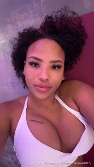 Woman with curly hair in white top, tattoo on chest, indoor setting.