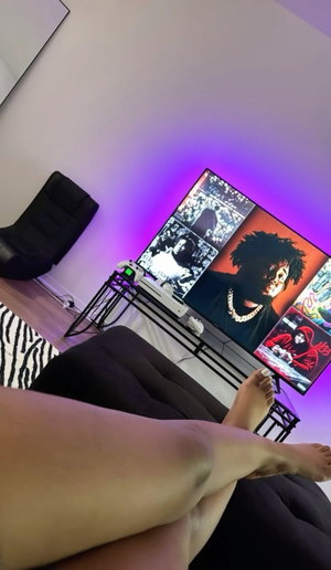 Person with bare legs and feet sitting on a black couch in a room with purple lighting and a large