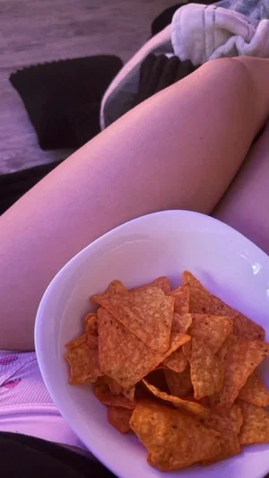 Person with bare legs holding a bowl of chips, wearing pink underwear, indoor setting.