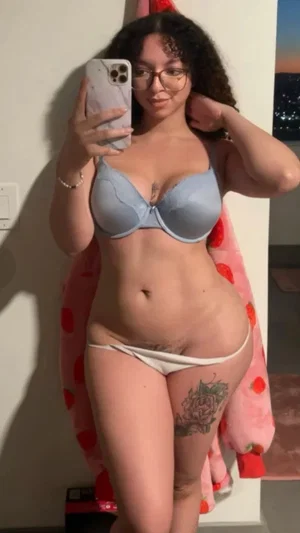 Woman in light blue bra and white underwear, standing indoors with curly hair and glasses.