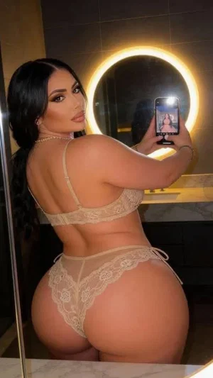 Woman in beige lace lingerie taking mirror selfie in bathroom.