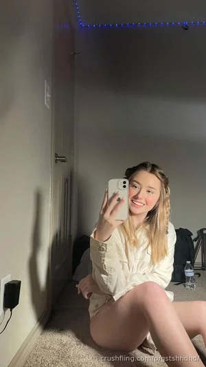Woman with blonde hair in a light-colored shirt sitting on floor, taking mirror selfie in room with