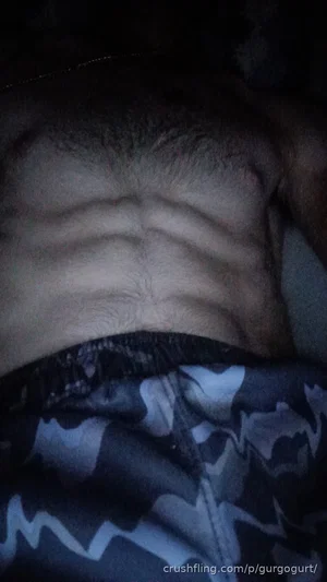 Man with defined abs wearing camouflage pants, lying down in dim light.