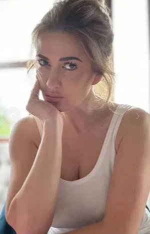 Woman with blonde hair in white tank top posing indoors.