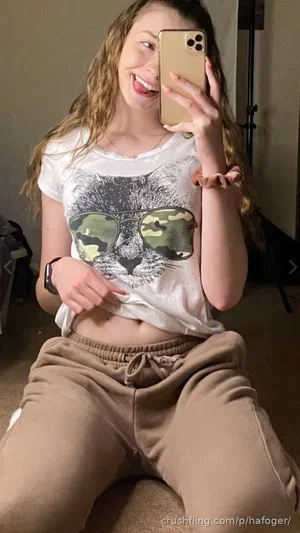 Woman with wavy hair in a cat graphic tee and sweatpants, sitting indoors taking a mirror selfie.