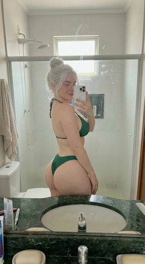 Woman with silver hair in green bikini taking mirror selfie in bathroom.