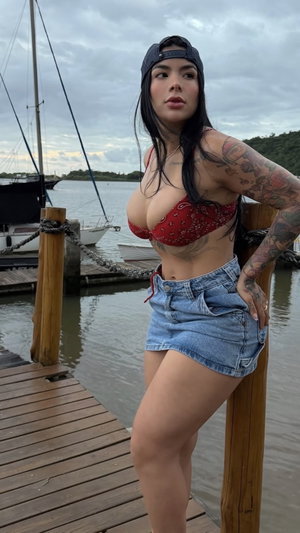 Tattooed woman in red top and denim shorts posing on dock.