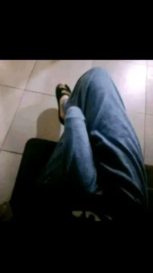 Person in blue pants and sandals sitting on a black chair on tiled floor.