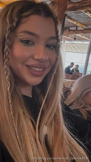 Woman with long blonde braided hair, wearing black top, smiling in a casual indoor setting.