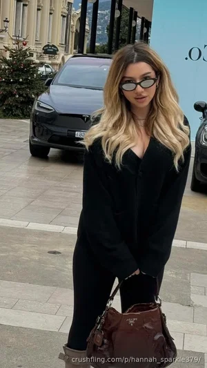 Woman with blonde hair in black outfit and sunglasses standing outdoors near cars.