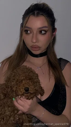 Woman with long hair holding a small dog, wearing a black mesh top and choker indoors.