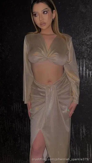 Woman in beige sheer dress with long sleeves and high slit, standing against dark textured wall.