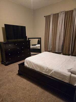 Bedroom with dark wood bed, dresser, TV and beige curtains.