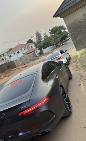 Two luxury cars parked on a dirt road near residential houses.