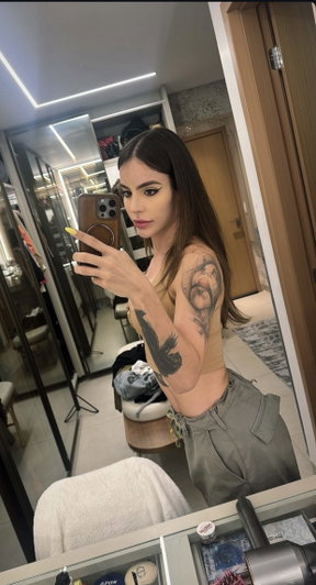 Woman with tattoos in beige top and cargo pants takes mirror selfie in modern room.