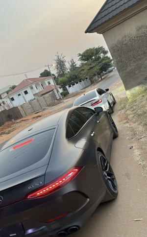 Two luxury cars parked on a dirt road near a house in a residential area.