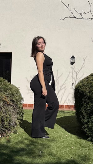 Woman in black one-shoulder jumpsuit posing outdoors on grass near a white wall.
