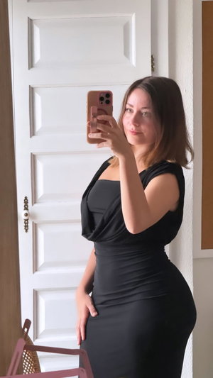 Woman in black dress taking mirror selfie indoors near white door.