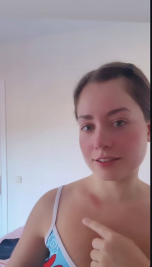 Woman in light blue top pointing at chest, indoor setting.