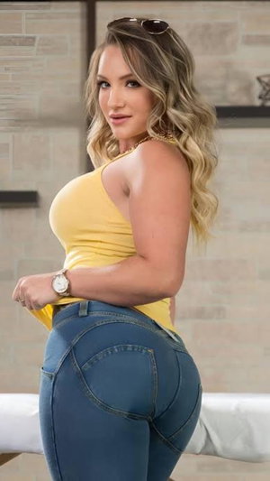 Woman with wavy blonde hair in yellow top and jeans, posing indoors.