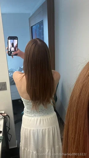 Woman with long brown hair in white dress taking mirror selfie in bedroom.