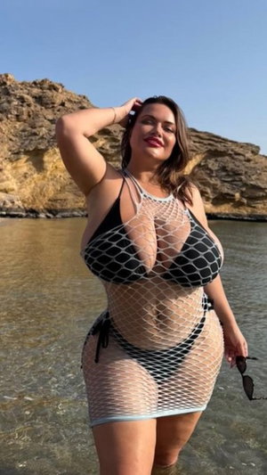 Woman in fishnet outfit standing in shallow water near rocky shore.