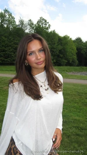 Woman with long brown hair in white off-shoulder top standing in grassy outdoor area.