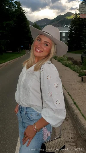 Blonde woman in white shirt and jeans with cowboy hat, standing outdoors near mountains.