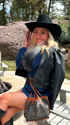 Blonde woman in black hat and leather jacket, sitting outdoors on patio with rocky background.