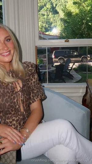 Blonde woman in leopard print top and white pants sitting indoors near window with outdoor view.