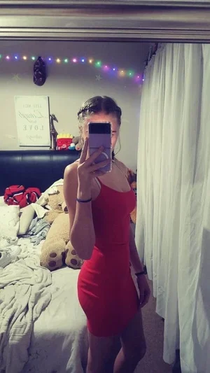 Woman in red dress taking mirror selfie in bedroom with bed and string lights.