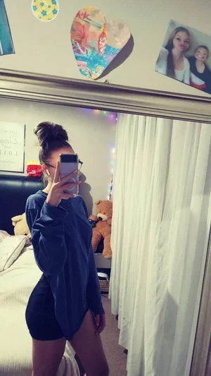 Woman in dark sweatshirt and shorts takes mirror selfie in bedroom with bed and stuffed animals
