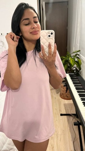 Woman in pink oversized t-shirt taking mirror selfie indoors, dark hair, keyboard in background.