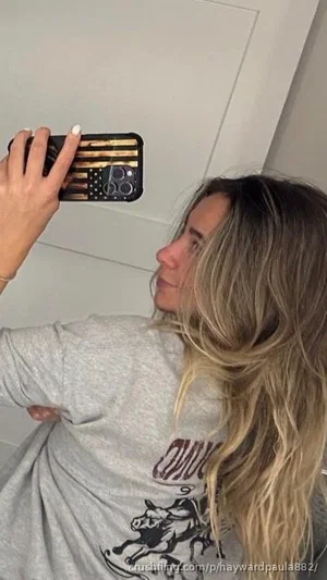 Woman with long blonde hair in gray t-shirt taking mirror selfie indoors.