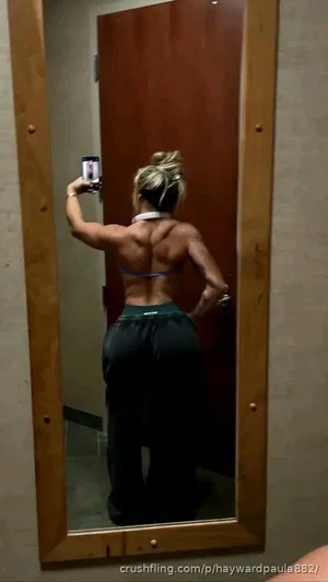 Woman with blonde hair in bun, wearing blue top and dark pants, taking mirror selfie in indoor