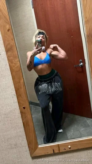 Woman in blue sports bra and black sweatpants flexing bicep in mirror selfie.