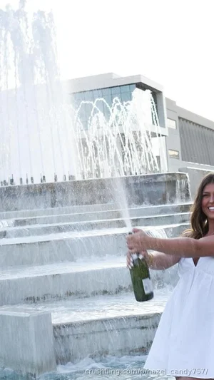 Woman in white dress holding champagne bottle near fountain, smiling.