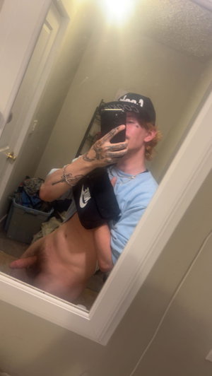 Person with tattoos and red hair in a mirror selfie, wearing a cap and partially unbuttoned shirt