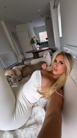 Buxom blonde in white ribbed bodysuit on couch indoors.